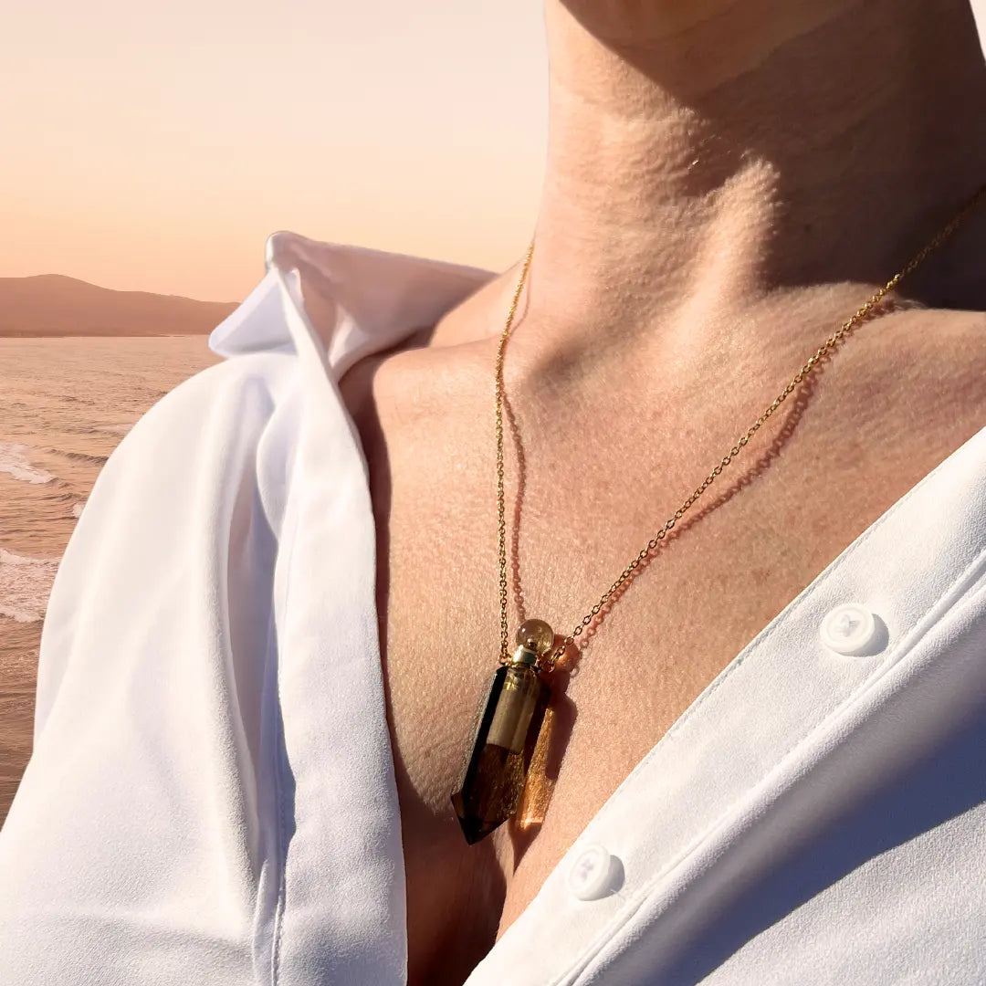 A person is adorned in a white shirt and an Ascention Beauty Crystal Perfume Amulet Necklace (Reward) featuring a brown pendant. The background showcases a tranquil beach scene at sunset with soft waves and distant hills, highlighting the natural crystal's charm.