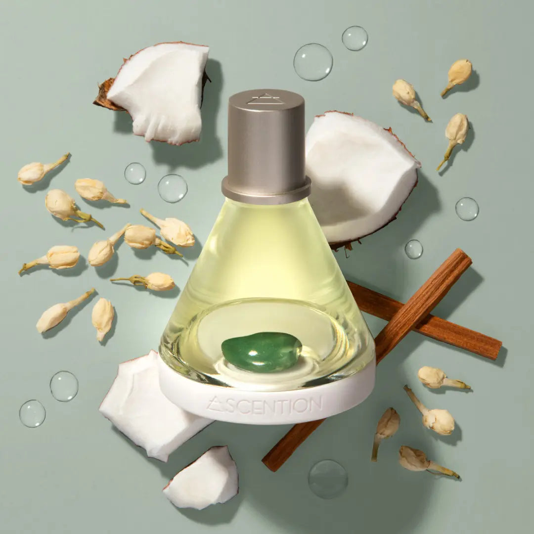 A bottle from Ascention Beauty's Powerful Scent Pairings collection, featuring a green gem in the base, is artfully surrounded by pieces of coconut, jasmine flowers, and cinnamon sticks—ideal for fragrance layering. This scent combination uplifts your mind and is elegantly displayed on a light green background with delicate droplets.