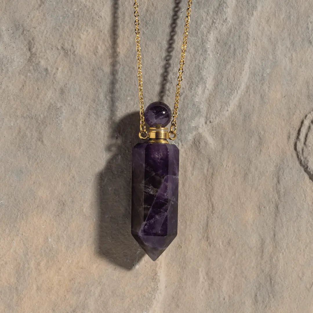 The Crystal Perfume Amulet Necklace (Reward) by Ascention Beauty effortlessly showcases a deep purple natural crystal in a hexagonal prism shape, accented with a round stone, and gracefully suspended from an 18K gold-plated chain. Its elegant design stands out against a textured stone background, casting a gentle shadow.