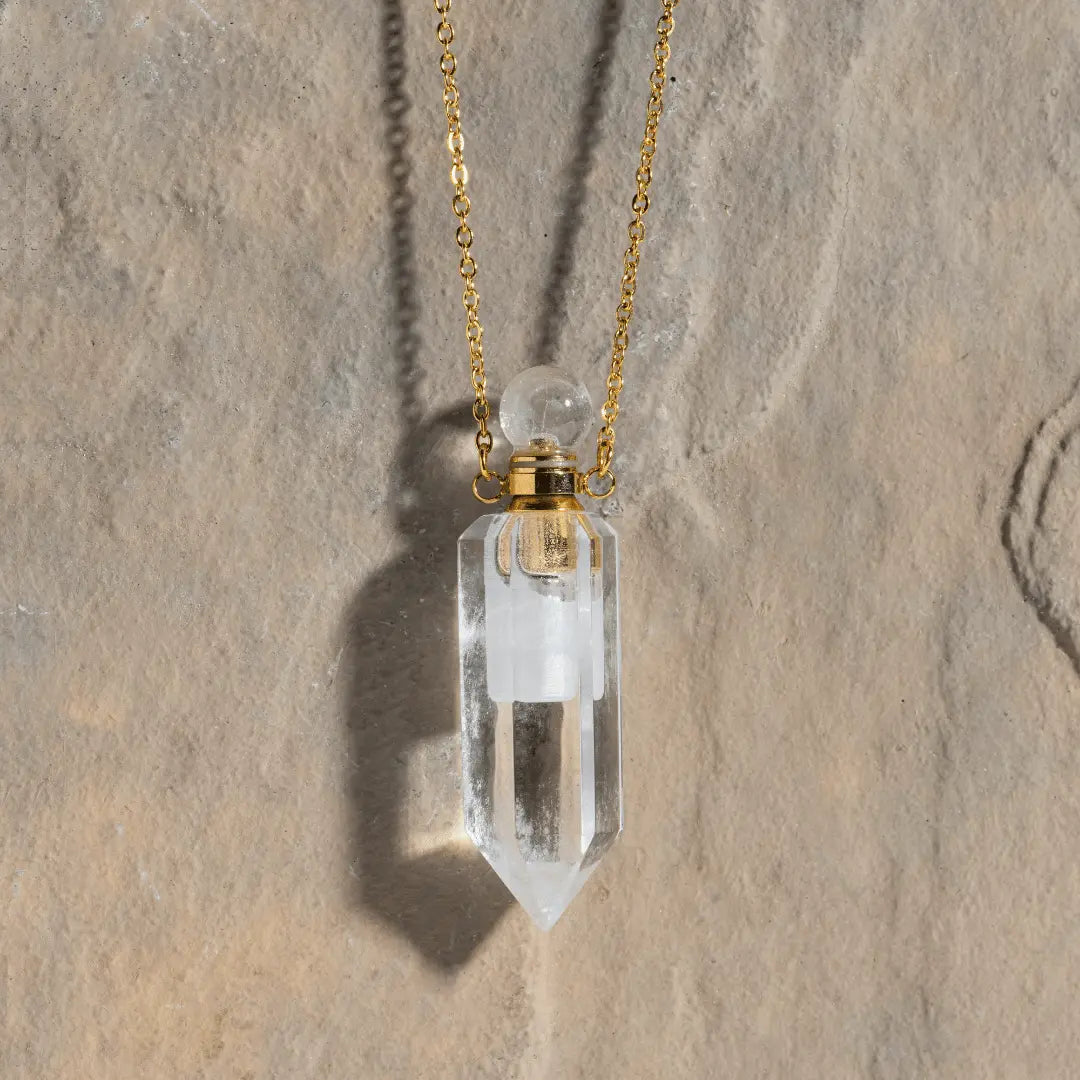 The Crystal Perfume Amulet Necklace (Reward) by Ascention Beauty - Wellness Clean Cruelty-Free Vegan Perfume features an 18K gold-plated chain that drapes elegantly against a textured stone backdrop. The pendant is crafted from a clear, faceted natural crystal with a spherical top, secured by a delicate gold cap. The light casts intriguing shadows from the pendant onto the stone surface.