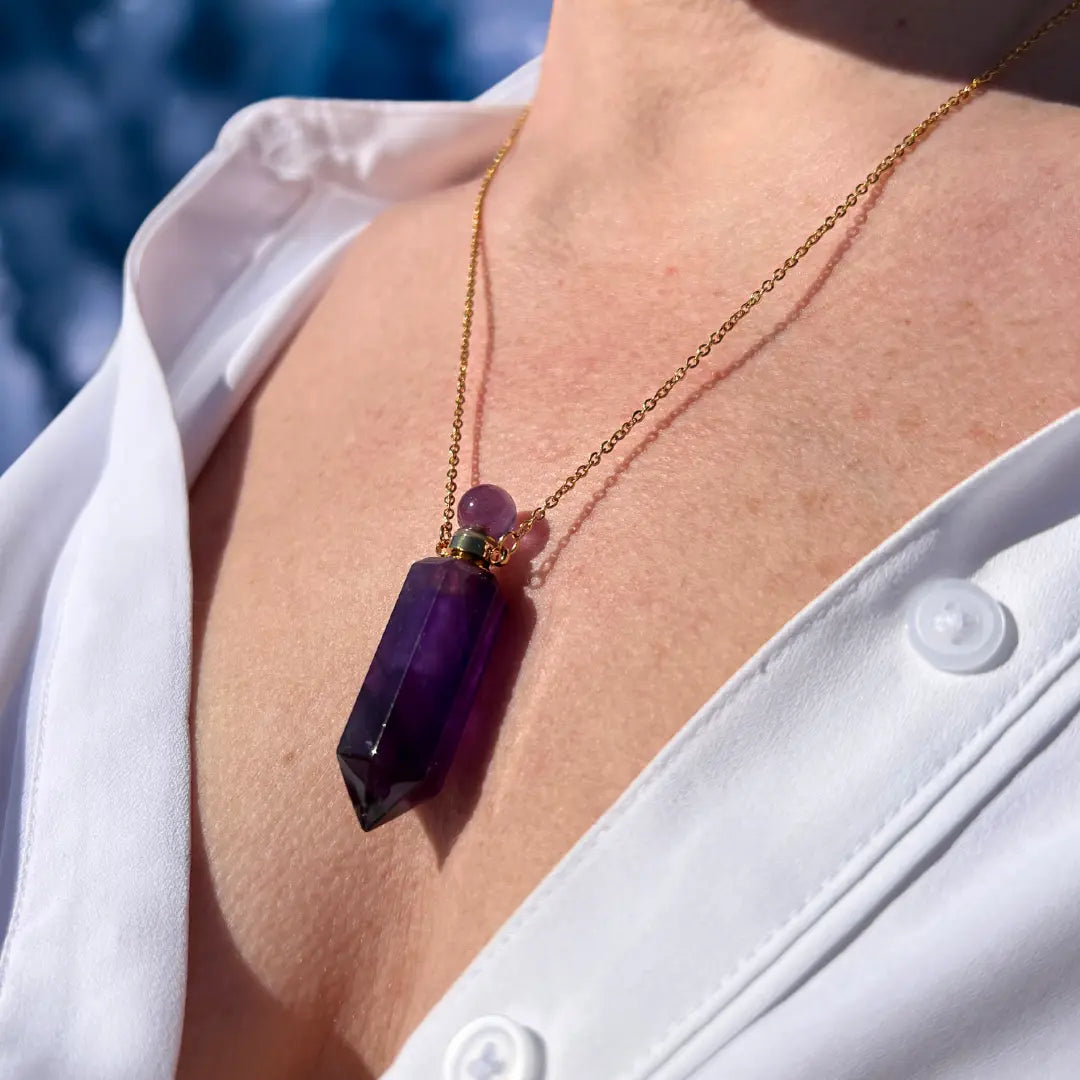 A close-up shows an individual in a white shirt adorned with the Crystal Perfume Amulet Necklace (Reward) by Ascention Beauty, a wellness brand known for its clean, cruelty-free, vegan perfumes. The necklace showcases a striking natural purple crystal suspended from an 18K gold-plated chain. The backdrop is softly blurred in shades of blue and white, evoking the serenity of a clear sky or water.