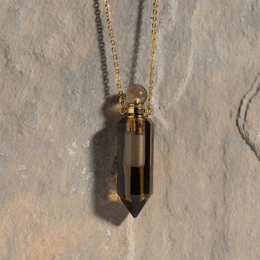 Hanging elegantly from a fine gold chain, the Crystal Perfume Amulet Necklace (Reward) showcases a pointed, translucent dark stone with a round top. Set against a textured stone background and casting a gentle shadow, this piece captures the spirit of ASCENTION Beauty's clean, cruelty-free vegan perfume.