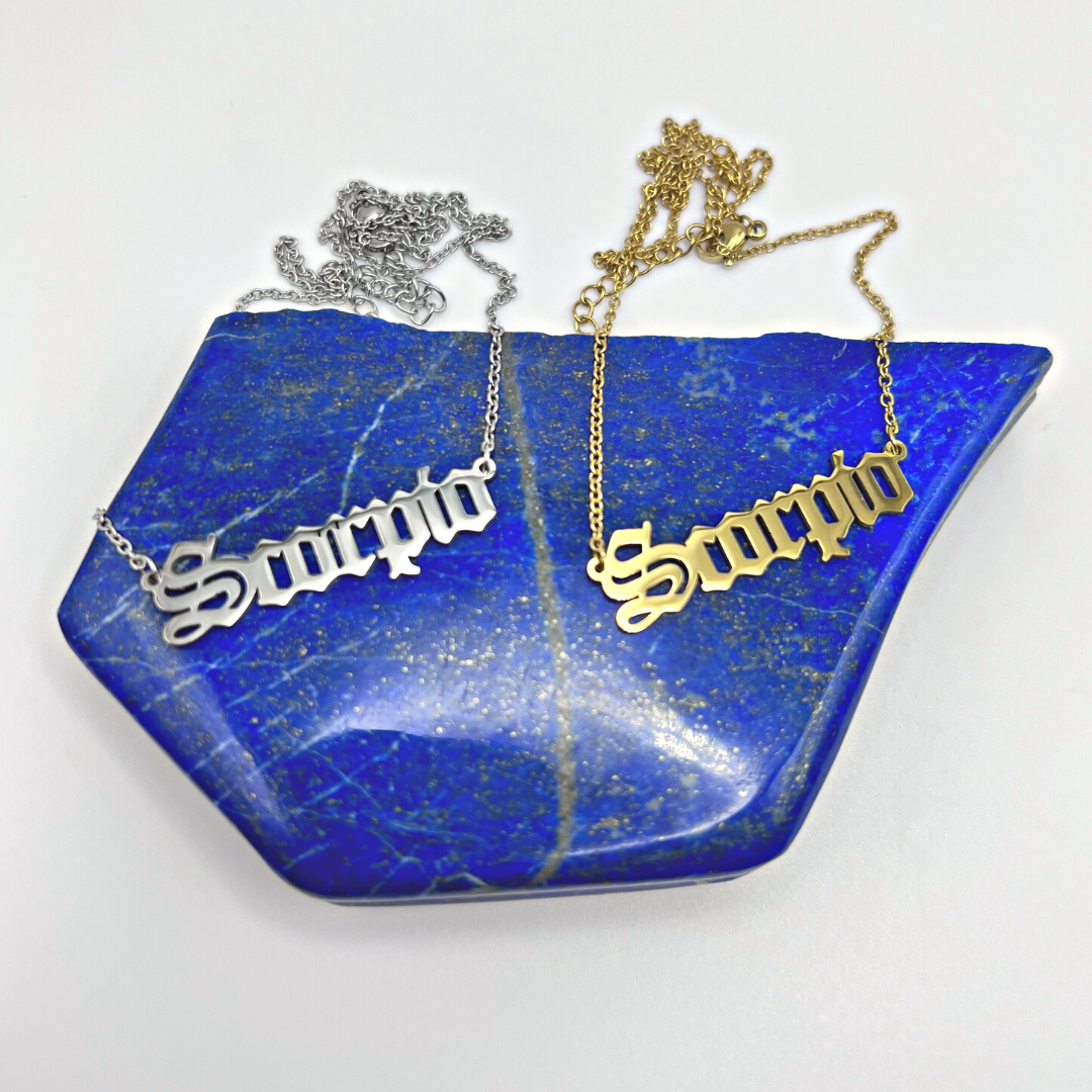 Two Zodiac Necklaces from Ascention Beauty, featuring Scorpio pendants in gold and silver, are elegantly displayed on a vibrant blue stone accented with subtle gold speckles, beautifully capturing the cosmic essence of celestial identity.