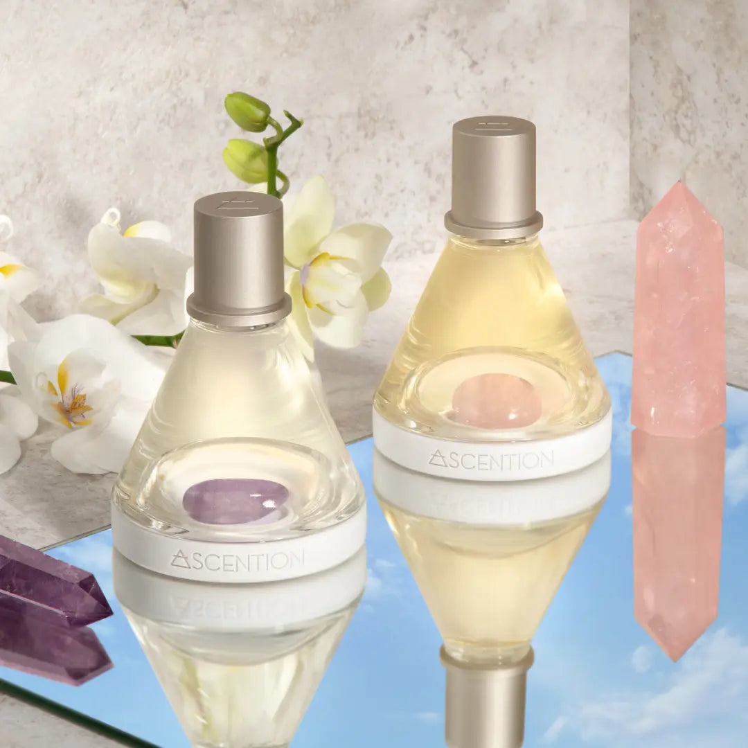 Two bottles of *Powerful Scent Pairings* from Ascention Beauty, featuring silver caps, rest gracefully on a reflective surface adorned with white orchids and purple crystals. A pink crystal stands upright beside them. This tranquil scene mirrors a blue sky with clouds, encouraging you to explore scent combinations that elevate your mind through clean, cruelty-free vegan perfumes.