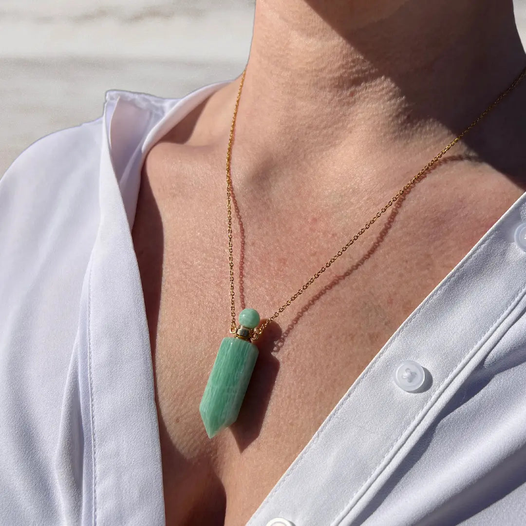 Close-up of a person wearing a white shirt and the Crystal Perfume Amulet Necklace (Reward) by Ascention Beauty, showcasing a green natural crystal pendant on an 18K gold-plated chain. The sunlight beautifully highlights the texture of both the crystal and the shirt.