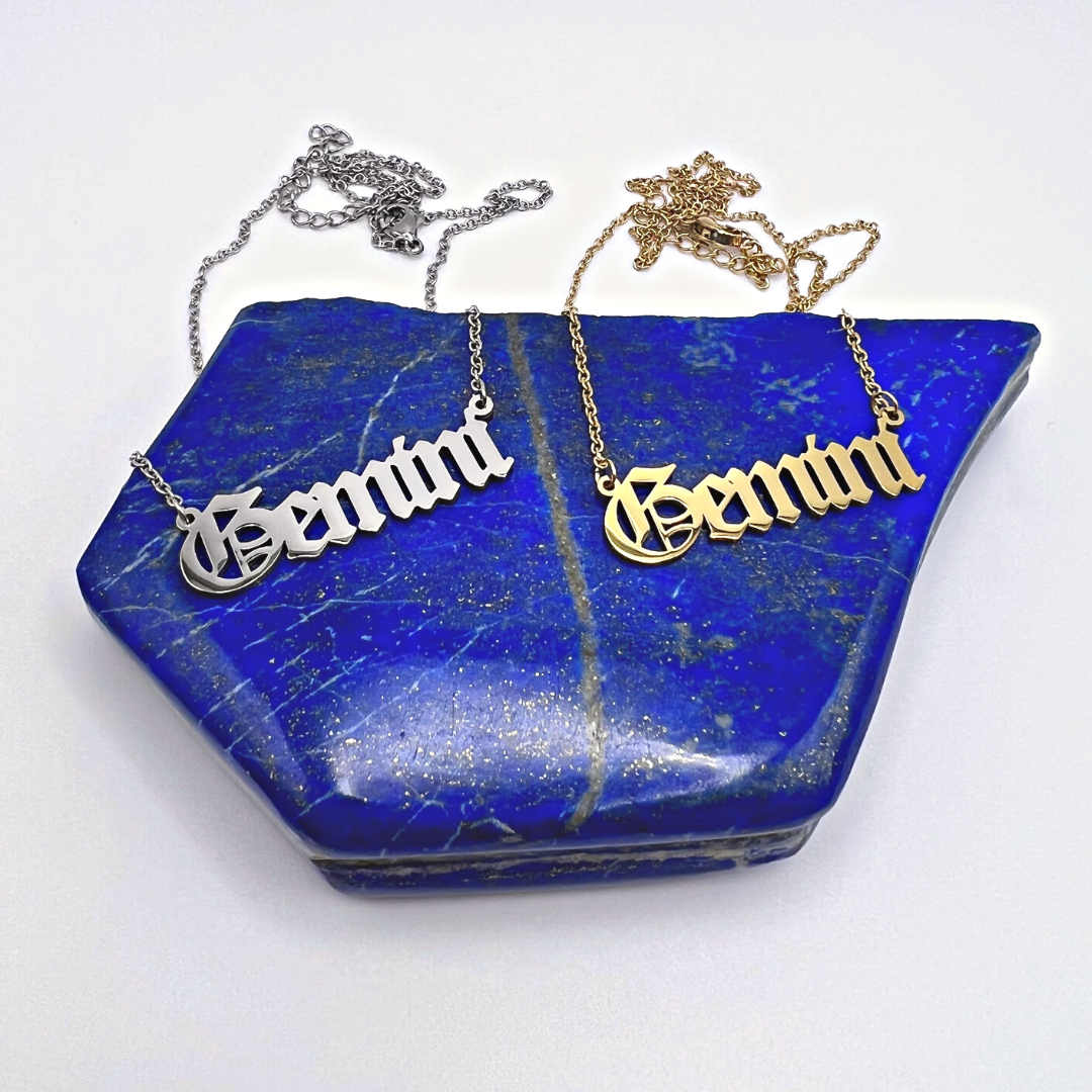 Two metallic Zodiac Necklaces by Ascention Beauty elegantly display the word "Gemini" on a slab of blue, gold-speckled stone. One necklace is in silver and the other in gold, both featuring cursive script that beautifully encapsulates your cosmic essence.