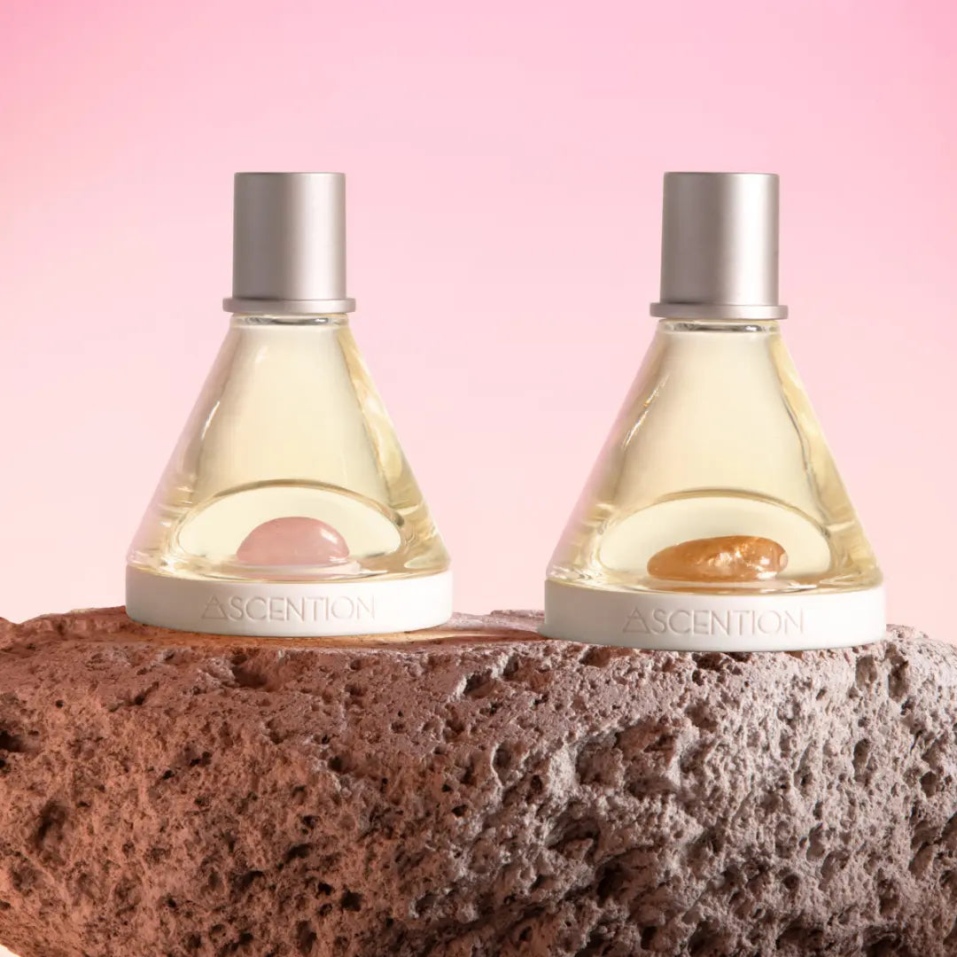 Two triangular bottles from the Powerful Scent Pairings collection by Ascention Beauty, each adorned with a silver cap and gemstone base—one pink and one amber—are elegantly arranged on a textured rock. These cruelty-free, vegan perfumes provide the perfect opportunity to explore unique scent combinations, against a backdrop of soft pink gradient tones.