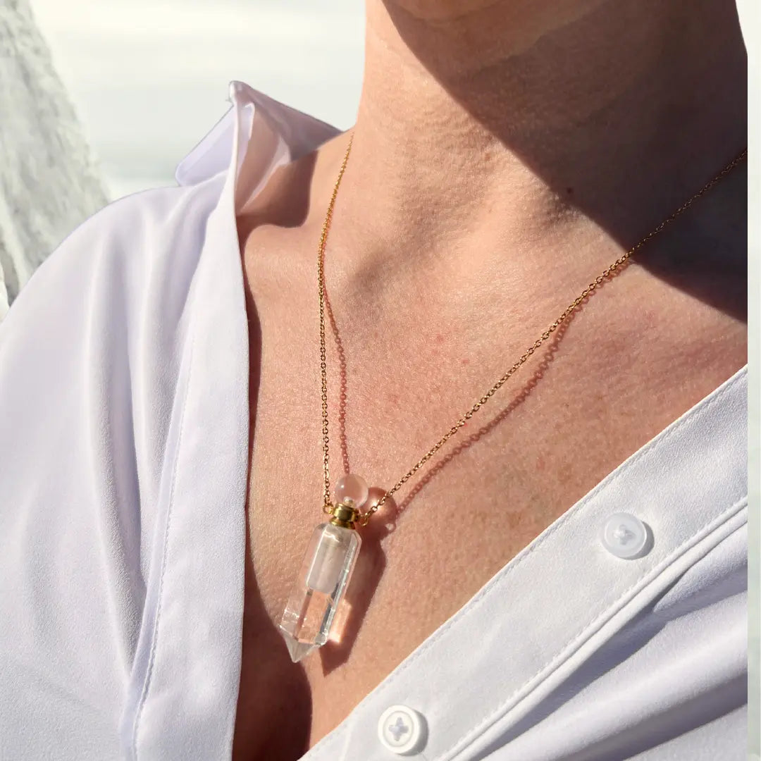 An individual wearing a white shirt displays an 18K gold-plated chain featuring the Crystal Perfume Amulet Necklace (Reward) by Ascention Beauty - Wellness Clean Cruelty-Free Vegan Perfume. The sunlight accentuates the natural crystal's details against their skin.