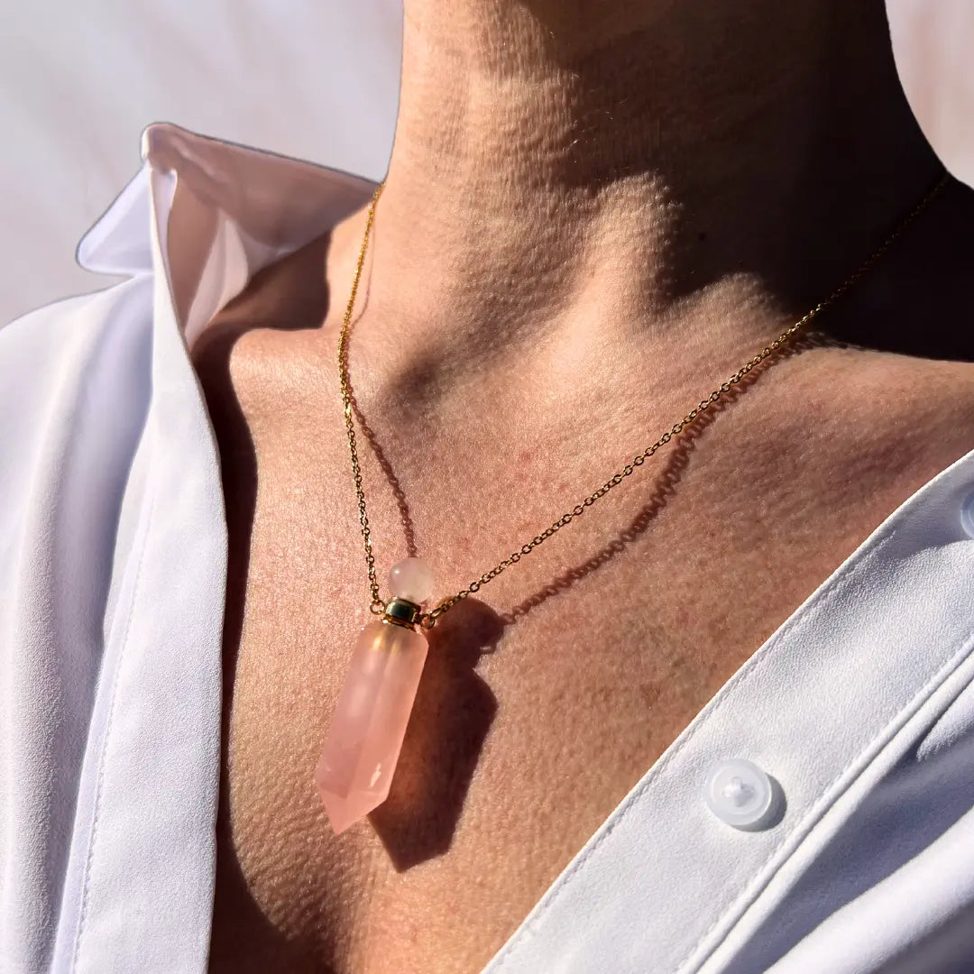 A close-up of a person wearing an open white shirt showcases the Crystal Perfume Amulet Necklace from Ascention Beauty, featuring a pink natural crystal pendant on an 18K gold-plated chain. The sunlight softly casts a shadow of this elegant piece on their skin.