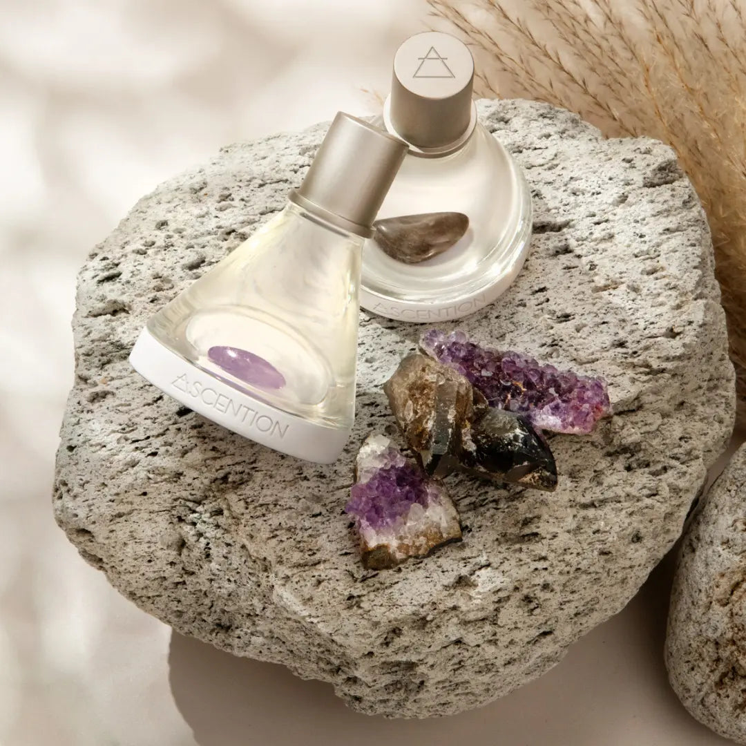 Two bottles from the "Powerful Scent Pairings" collection by Ascention Beauty are displayed on a textured stone, accentuated with amethyst crystals, encouraging fragrance layering. Their elegant design features one bottle upright and the other tilted, creating a serene and luxurious presentation to uplift your senses.