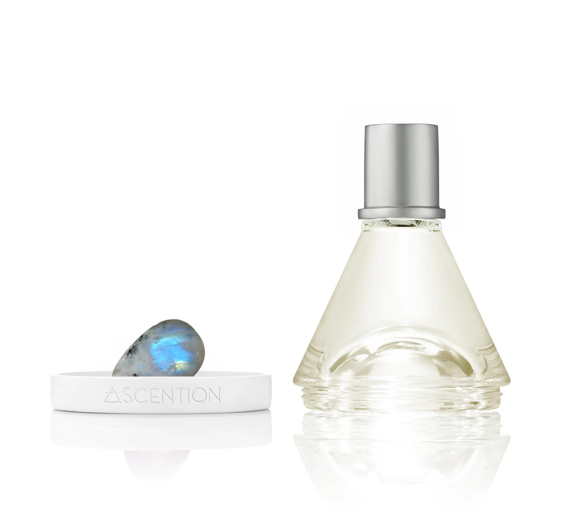 A triangular glass bottle of the *NEW* Ascent to Enchant With Rainbow Moonstone Perfume, featuring a silver cap, sits beside a white dish holding a small, iridescent Rainbow Moonstone. The dish is engraved with ASCENTION BEAUTY CO. The white background accentuates the elegance of these enchanting items, perfectly suited for a dark feminine goddess.