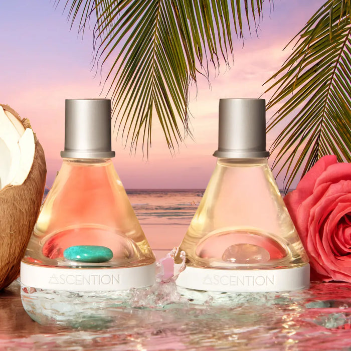 On a reflective surface against the backdrop of a tropical sunset with palm leaves, two bottles of Ascention Beauty's Powerful Scent Pairings perfume are elegantly positioned. A pink rose, coconut, and turquoise stones enhance the setting as waves softly reach the base of the bottles. Immerse yourself in these unique scent combinations designed to elevate your mind and transport you to paradise, all while embracing wellness with clean, cruelty-free vegan products.